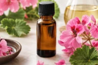 Amber bottle of rose geranium essential oil sitting amongst flowers and a flask of oil